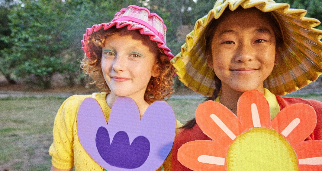 Two young girls in handcrafted flower costumes and hats smile in an outdoor setting with soft, diffused light, trees, and greenery.