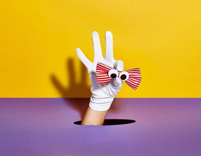 Hand in white glove emerges from purple hole displaying googly-eyed striped bow tie against yellow backdrop, studio lighting.