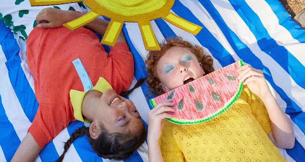 Two smiling children lying on a striped blanket with a paper sun and watermelon, enjoying a sunny day in a playful, colorful scene.