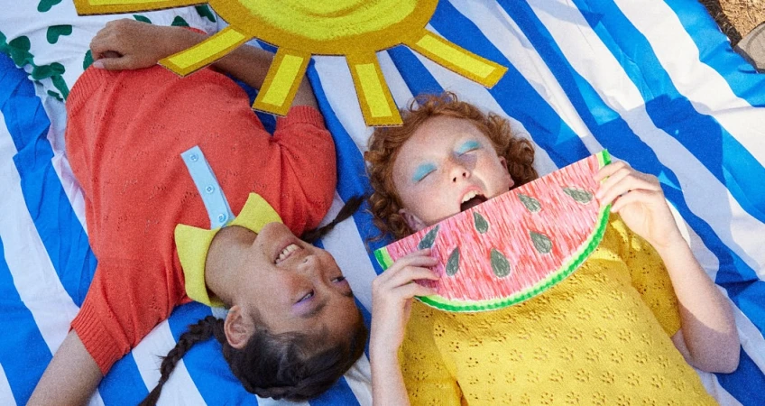 Two smiling children lying on a striped blanket with a paper sun and watermelon, enjoying a sunny day in a playful, colorful scene.