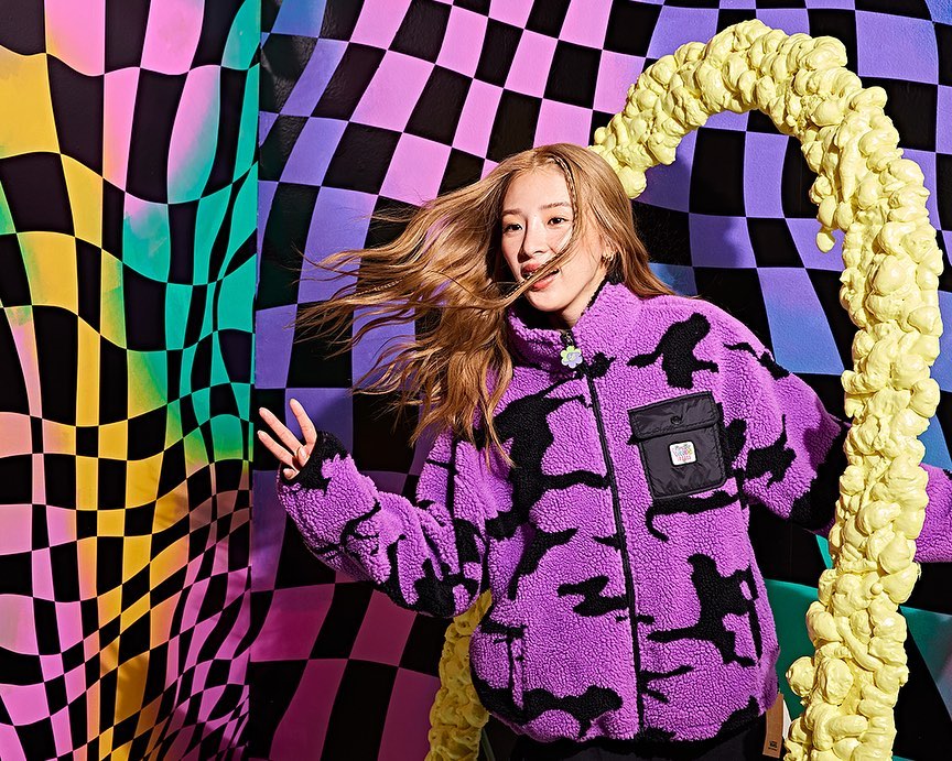 Portrait of a young adult female with long wavy hair smiling in a purple jacket against a colorful checkerboard pattern background.