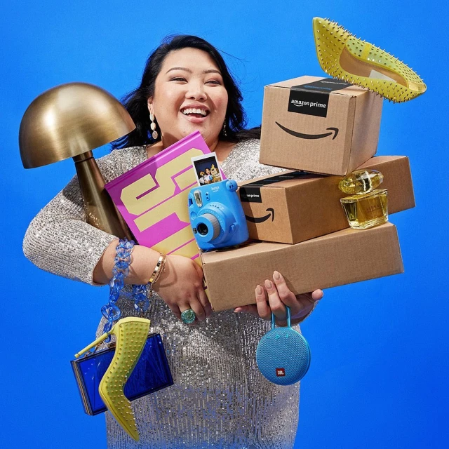 Smiling woman in a sequined dress poses with a stack of Amazon shipping boxes, camera, perfume bottle, speaker, purse, and a lamp.