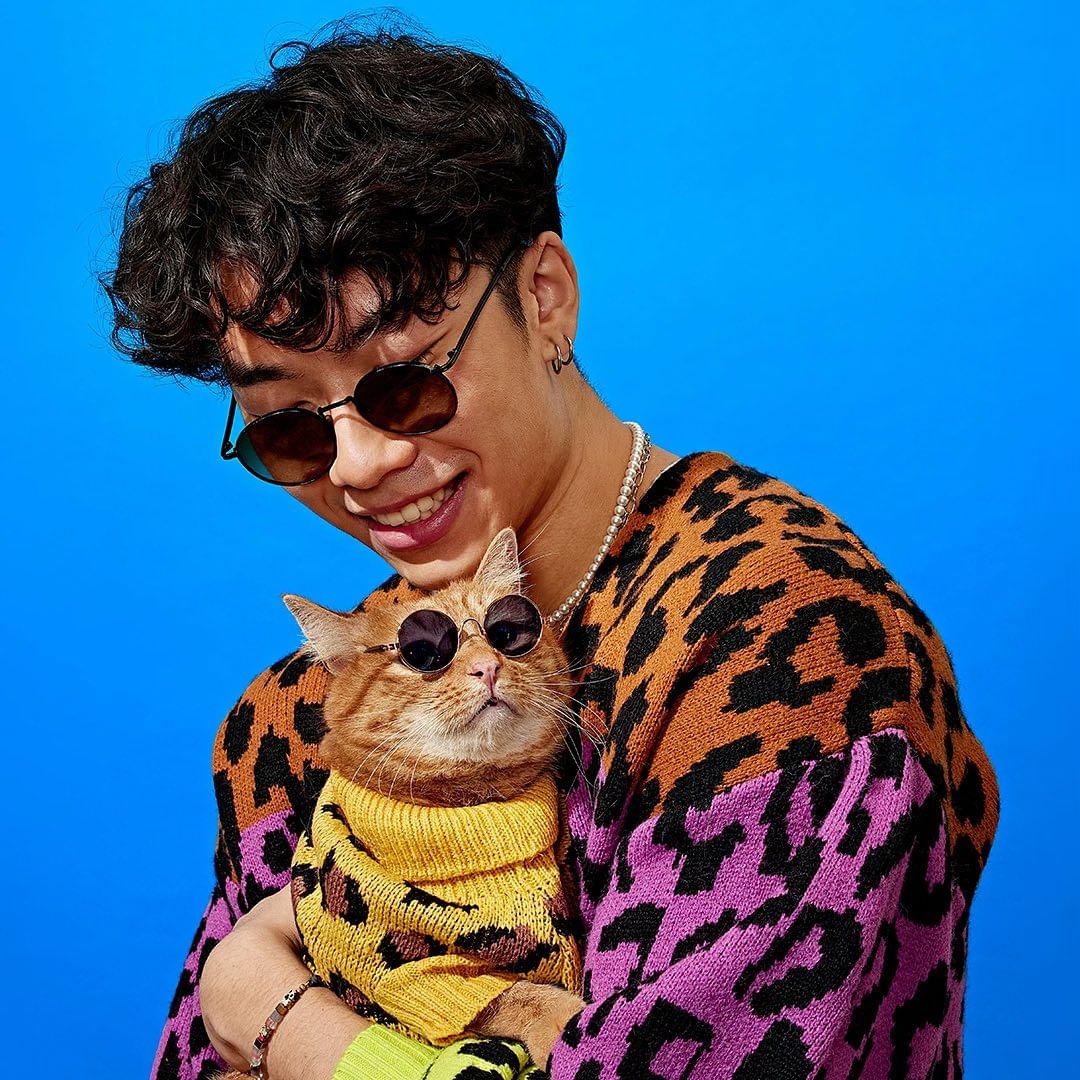 Stylish man smiling, holding a ginger cat wearing a yellow sweater and sunglasses against a bright blue backdrop, fun fashion.