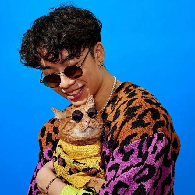 Stylish man smiling, holding a ginger cat wearing a yellow sweater and sunglasses against a bright blue backdrop, fun fashion.