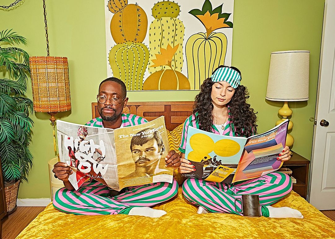Stylish couple in striped pajamas sits on a yellow bed reading magazines, with a vibrant cactus painting, retro lamp, and green wall background.
