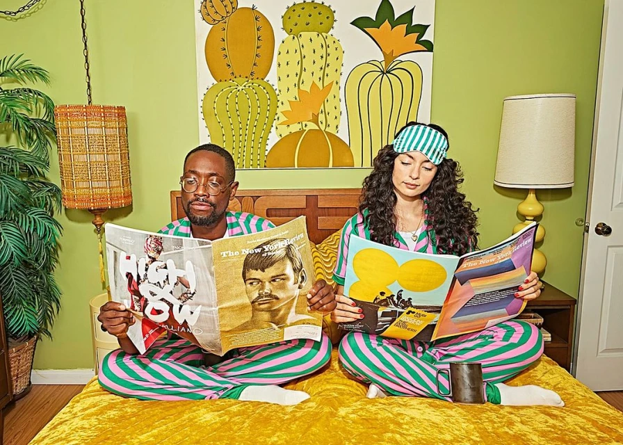 Stylish couple in striped pajamas sits on a yellow bed reading magazines, with a vibrant cactus painting, retro lamp, and green wall background.