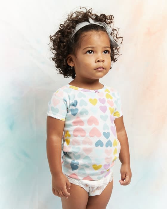 Portrait of an African-American toddler with curly hair, wearing a heart-print shirt and diaper, looking up against a painterly background.