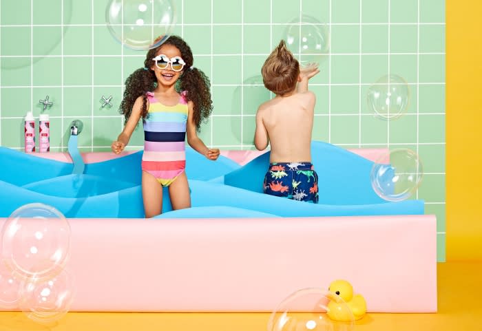 Two children, a girl and a boy, play in a pastel-colored tub filled with blue water, surrounded by bubbles and bright yellow background.