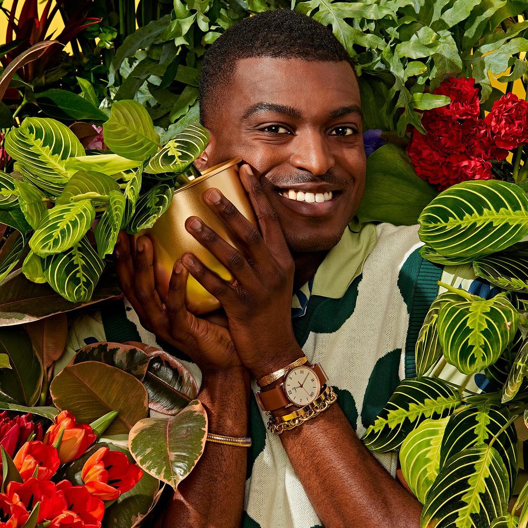 Smiling African-American man surrounded by plants and flowers, cradling a gold pot with leafy green plant. Close-up, indoor portrait.