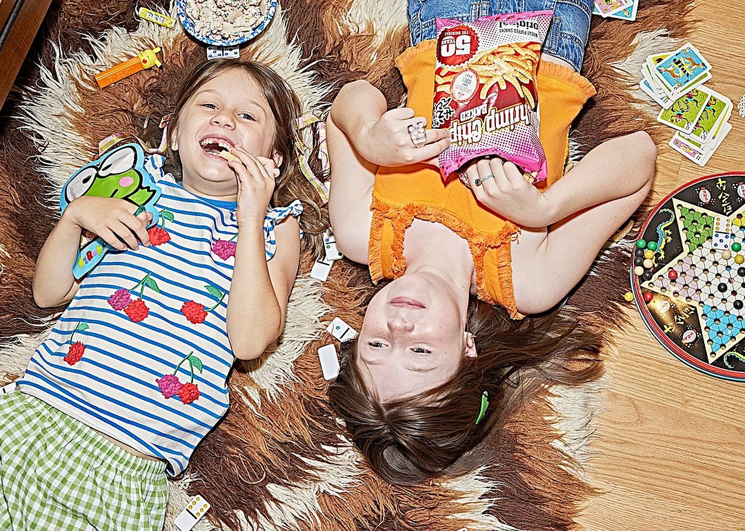 Two young girls lie on a fur rug, playing with board games, cards, and dominoes, with one girl holding a bag of chips in an indoor setting.