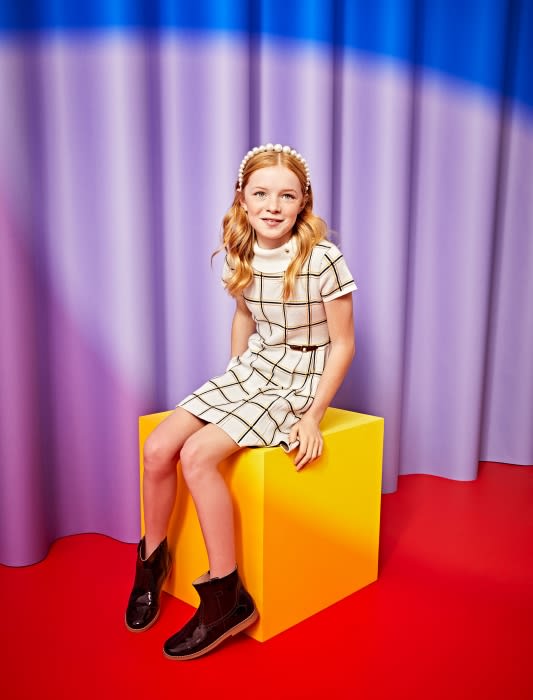 A young girl with red hair, wearing a white and black grid-patterned dress and patent black boots, sits on a yellow cube in a photo studio.