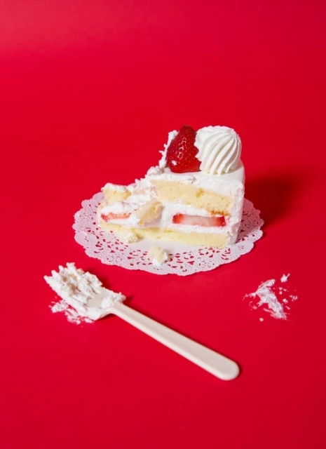 Slice of strawberry shortcake with whipped cream and a plastic fork set against a vibrant red background in a food photography studio setup.