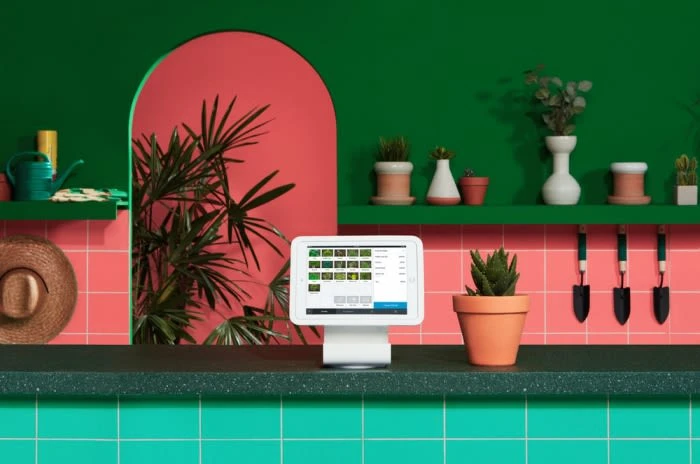 Still life features a tablet, potted succulents, watering can, and hat against a green and pink tile backdrop, indoor setting.