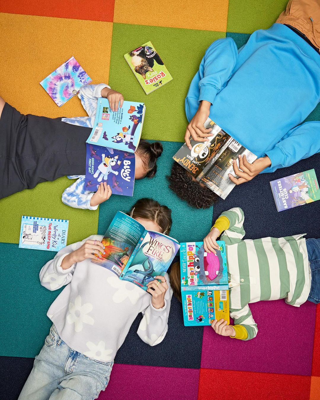 Overhead shot of four children lying on a colorful, square-patterned carpet, each holding and reading a different book. Reading activity.