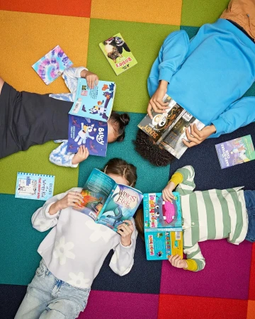 Overhead shot of four children lying on a colorful, square-patterned carpet, each holding and reading a different book. Reading activity.