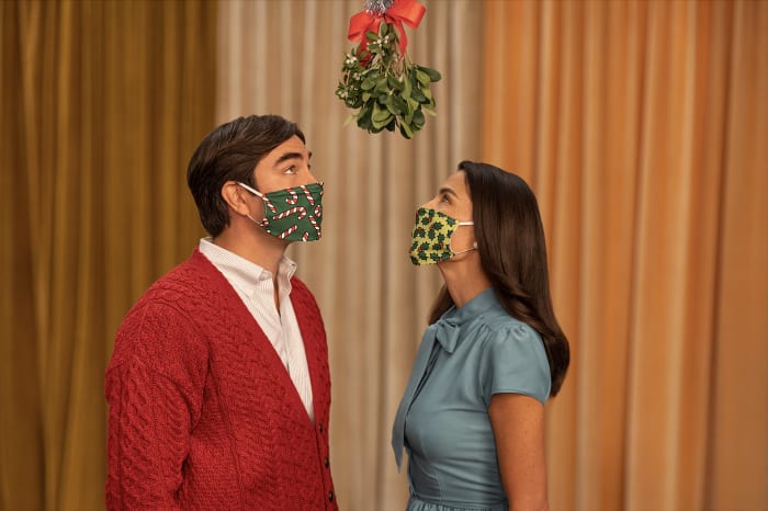 A couple wearing festive face masks looks up at mistletoe against a gold curtain backdrop, creating a holiday-themed scene with a focus on safety.