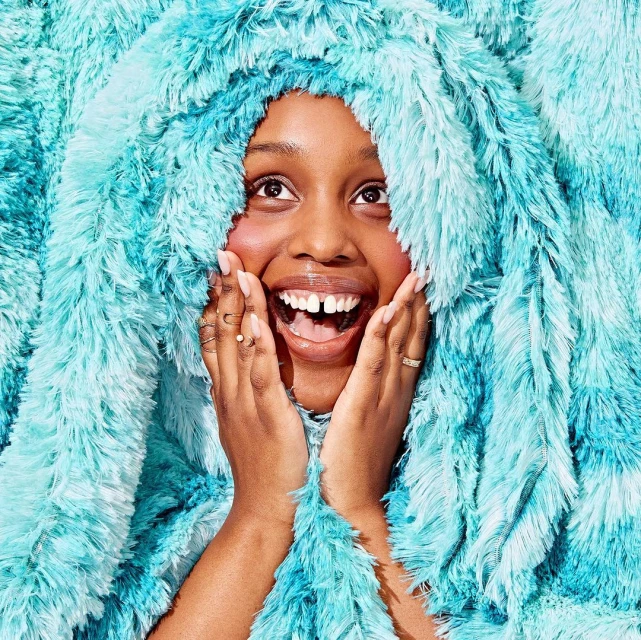 Smiling woman with hands on face, framed by a large, soft turquoise shag material. Bright lighting, portrait-style framing, indoor setting.