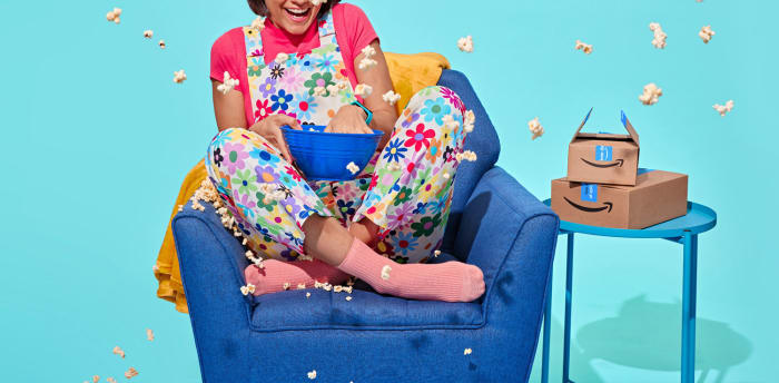 A woman sits in a blue chair throwing popcorn in the air, wearing colorful flower overalls, next to Amazon boxes, set against a blue backdrop.