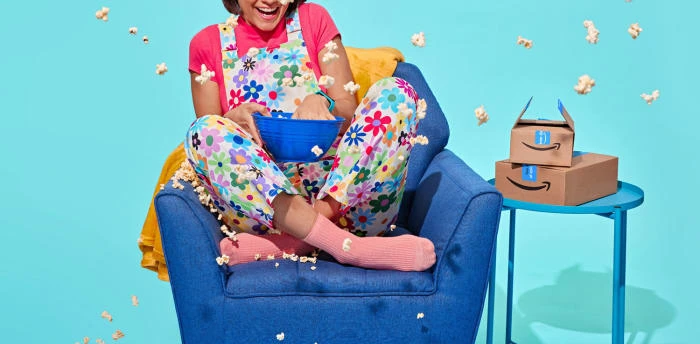 A woman sits in a blue chair throwing popcorn in the air, wearing colorful flower overalls, next to Amazon boxes, set against a blue backdrop.