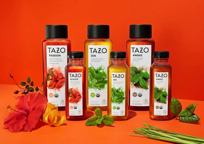Bottles of Tazo tea drinks are arranged on a bright orange surface with flowers and herbs, showcasing vibrant colors and fresh ingredients.