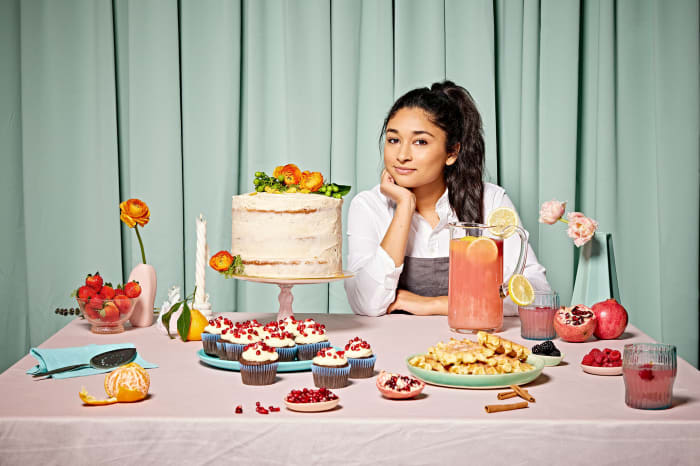 Adult female smiles behind a table laden with a cake, cupcakes, waffles, fruit, and drinks in front of a mint green backdrop.