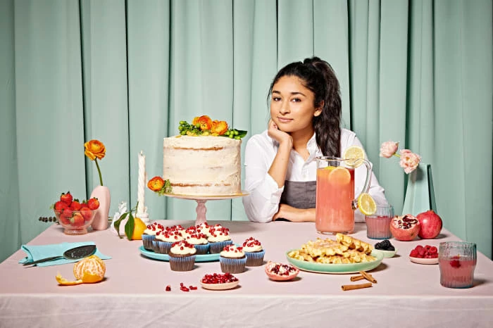 Adult female smiles behind a table laden with a cake, cupcakes, waffles, fruit, and drinks in front of a mint green backdrop.