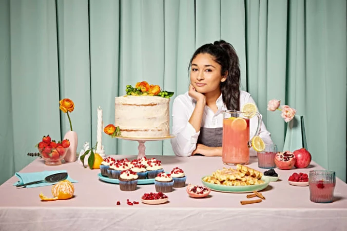 Adult female smiles behind a table laden with a cake, cupcakes, waffles, fruit, and drinks in front of a mint green backdrop.