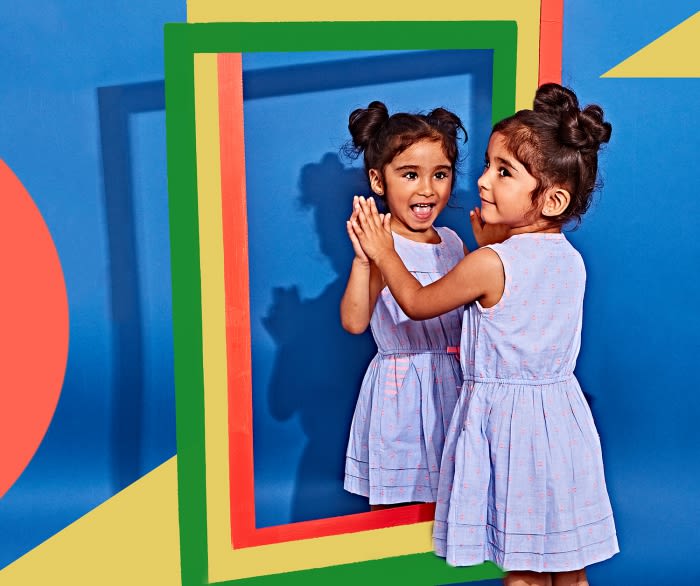 Young girl in light blue dress with pigtails smiles, reflected in frame; bright blue background with colorful geometric shapes.