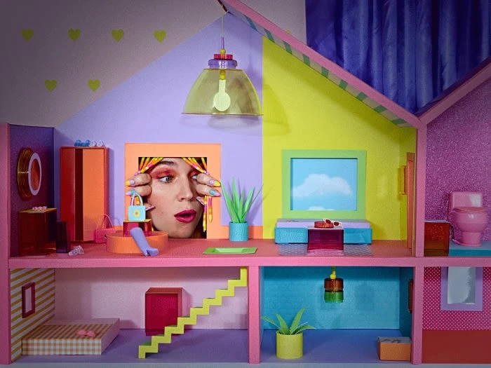 Surreal dollhouse interior with woman's face peering through a frame, colorful furniture, and playful decor in a whimsical setting.