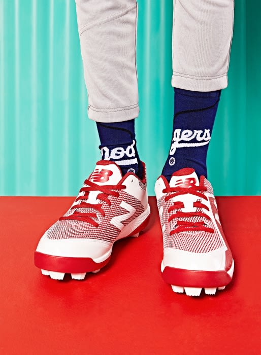 Close-up studio shot of red and white New Balance baseball cleats with navy blue socks on a red and teal background.