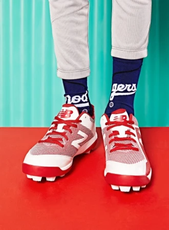 Close-up studio shot of red and white New Balance baseball cleats with navy blue socks on a red and teal background.