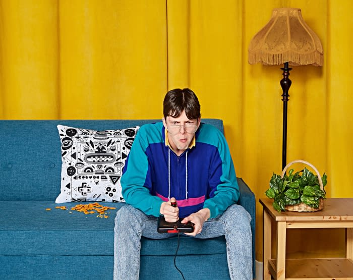Man in glasses plays video game on blue couch with chips and retro lamp against yellow backdrop indoors, looking focused on screen.