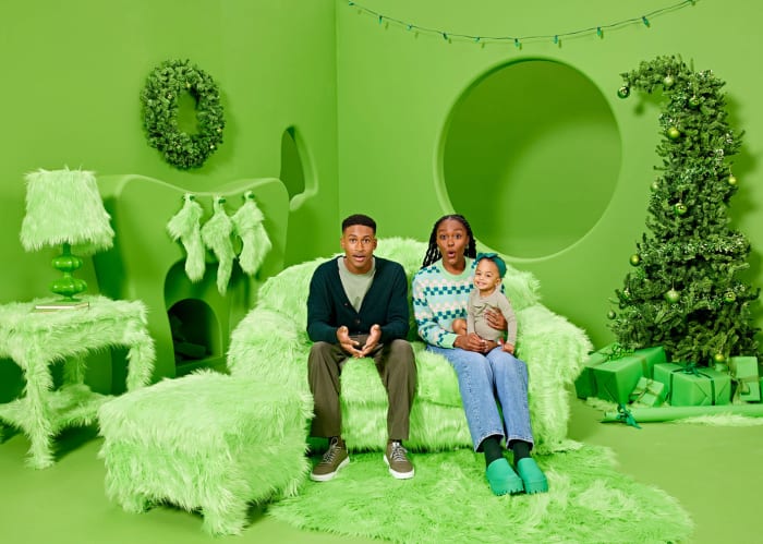 Family portrait in a green monochrome photo studio with parents and toddler daughter, seated on a fluffy green couch by a Christmas tree.