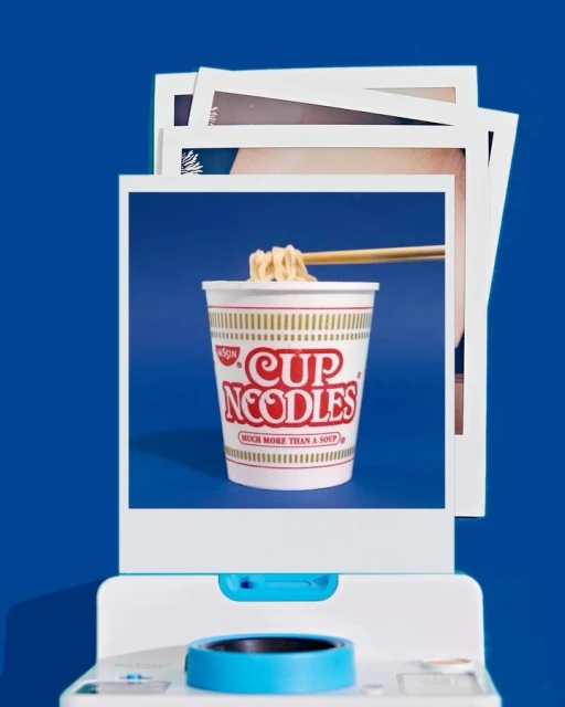 Cup Noodles instant ramen in cup with chopsticks, framed in a white Polaroid frame on a white printer against a blue background.