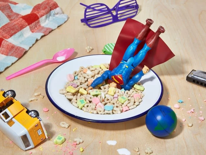 Superman action figure dives headfirst into a bowl of cereal with lucky charms, surrounded by toys on a wooden surface.