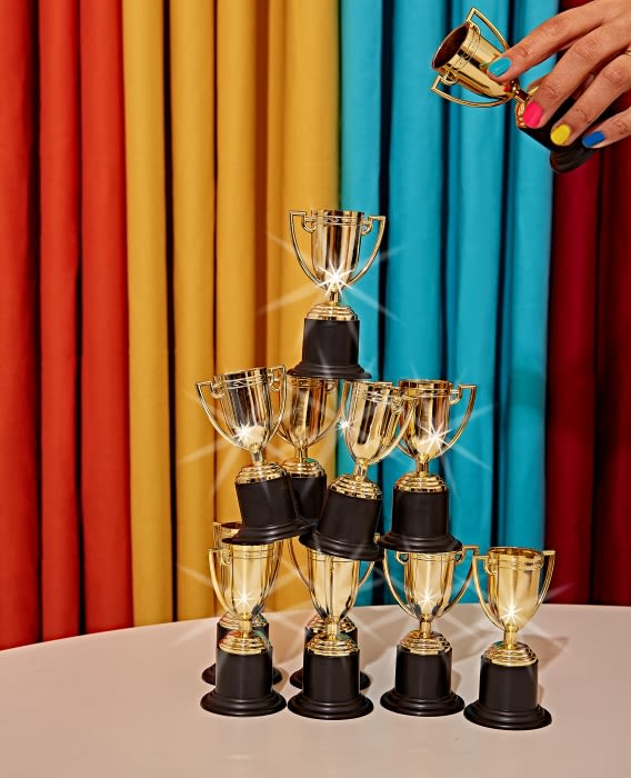 Stack of gold trophies on a white table, with a hand holding a trophy above, set against a colorful draped background.