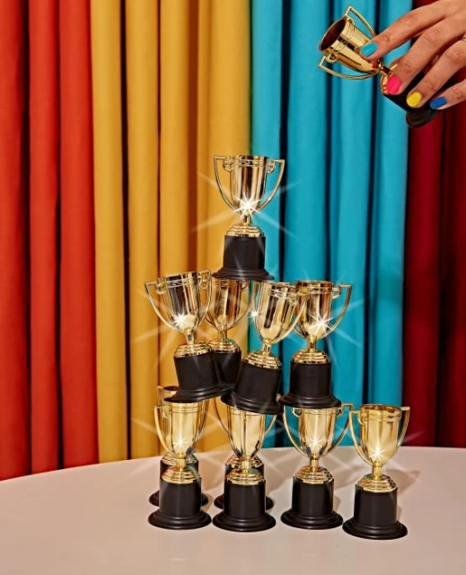 Stack of gold trophies on a white table, with a hand holding a trophy above, set against a colorful draped background.