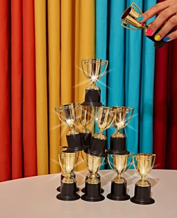 Stack of gold trophies on a white table, with a hand holding a trophy above, set against a colorful draped background.