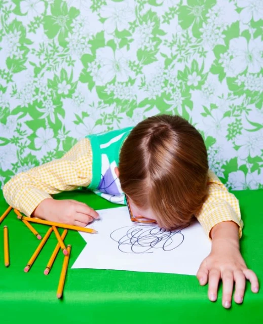 Child with eyeglasses rests head on a drawing, surrounded by pencils, on a green table against a floral patterned wallpaper.