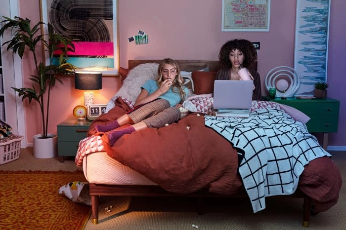 Two women relax on a bed in a bedroom, one eating popcorn and the other drinking from a cup while looking at a laptop.