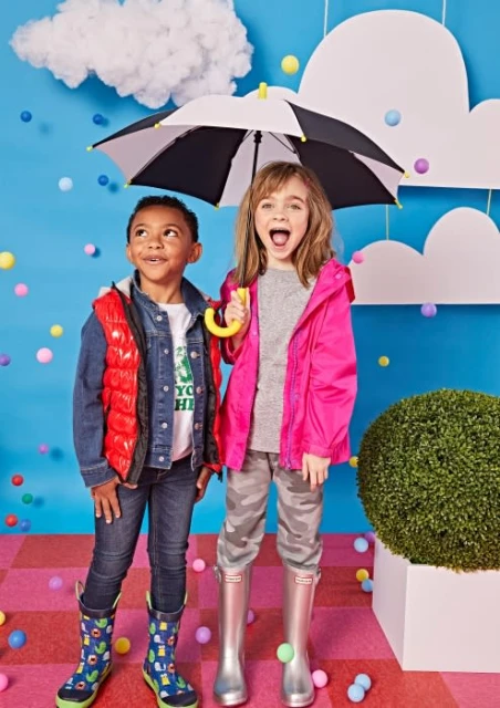 Two children, a boy and a girl, stand under an umbrella in a studio with a blue background, wearing rain boots and jackets.