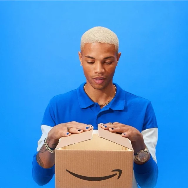 Young adult man with blonde hair opening a cardboard box with a curved arrow on a blue background. The man is wearing a polo shirt.