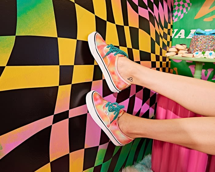 Legs and feet wearing tie-dye Vans slip-on sneakers are featured against an abstract checkered backdrop with cake and cookies.
