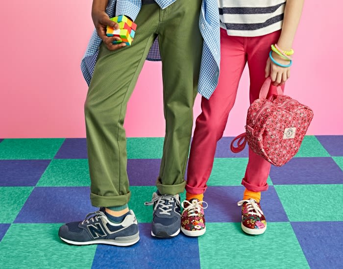 Two children stand on a blue and green checkered floor, wearing stylish pants, colorful socks, and sneakers; a backpack and Rubik's cube.