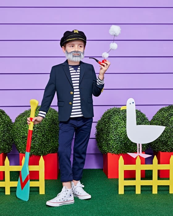 A young boy dressed in a sailor costume with a captain's hat, striped shirt, and navy blazer, holding a toy pipe and oar, outdoors.