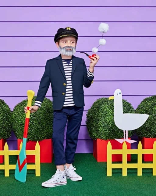 A young boy dressed in a sailor costume with a captain's hat, striped shirt, and navy blazer, holding a toy pipe and oar, outdoors.
