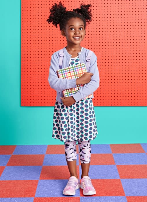 A young girl with curly hair, dressed in a polka-dot dress and lavender cardigan, holds a plaid book in front of a colorful background.