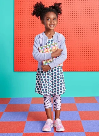 A young girl with curly hair, dressed in a polka-dot dress and lavender cardigan, holds a plaid book in front of a colorful background.