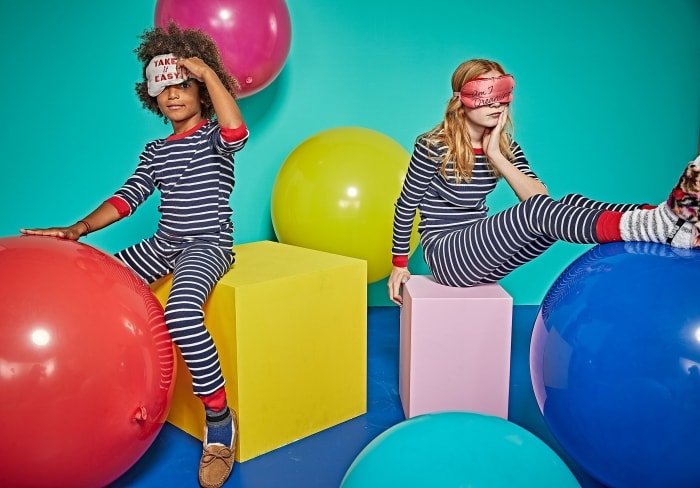 Two children in striped pajamas sit on oversized props in a brightly colored room. One wears a sleep mask, creating a playful, dreamy mood.