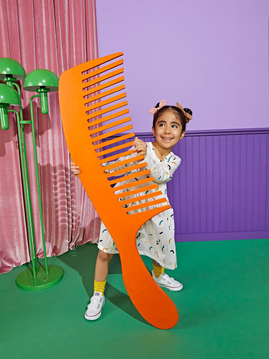 Young girl smiles while holding a large orange comb, standing indoors with a purple wall and green floor, adding a whimsical touch.